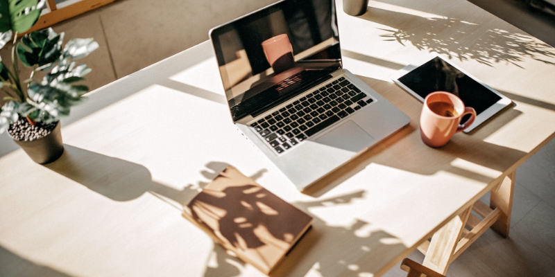Foto de estação de trabalho com notebook, tablet, xícara e vaso de planta em frente a uma janela simbolizando o trabalho remoto de equipe autogerenciável